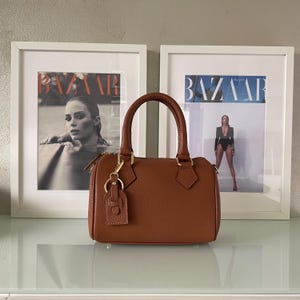May include: A brown, pebbled leather handbag with two top handles, a top zipper closure, and a matching leather luggage tag attached to a gold-toned metal ring on the front. The bag features a structured, rectangular shape with rounded corners.
