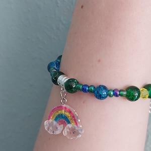 Puede incluir: Una pulsera de cuentas con un dije de arcoíris. La pulsera presenta cuentas verdes, azules y transparentes. El dije de arcoíris tiene un diseño colorido con arcos rojos, amarillos, azules y verdes, y nubes brillantes transparentes.