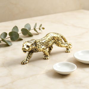 Könnte beinhalten: Goldfarbene Metall-Leopard-Figur mit gepunktetem Muster, in Gehposition. Zwei kleine weiße Schalen befinden sich im Hintergrund. Die Figur steht auf einer Marmoroberfläche.
