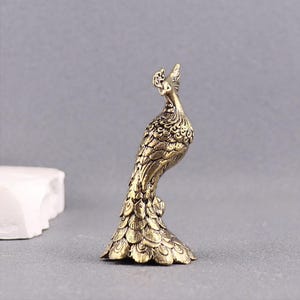 Puede incluir: Una peque&ntilde;a figura de pavo real en metal dorado con intrincados detalles de plumas en relieve en todo el cuerpo y una base en forma de abanico. El ave se muestra en posici&oacute;n erguida con la cabeza ligeramente girada hacia un lado.