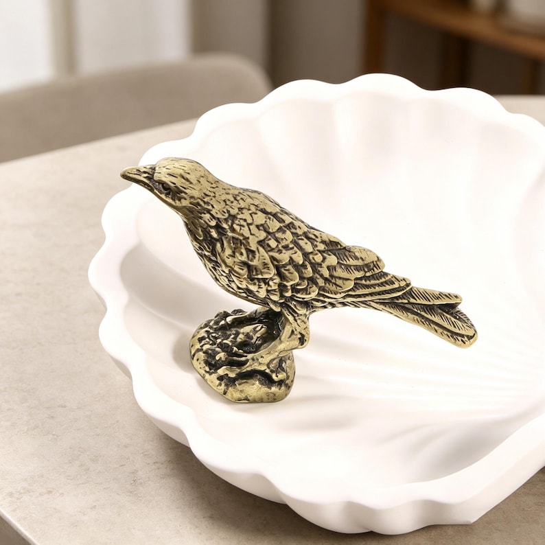May include: A detailed brass bird figurine perched on a textured base, set in a white, scalloped-edge dish. The bird has intricate feather detailing and a slightly aged appearance, suggesting a vintage or antique aesthetic. The dish provides a clean, contrasting backdrop.