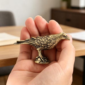 May include: A detailed brass raven figurine. The bird is perched on a small base, with intricate feather detailing. The raven is held in a person's hand, showcasing its size and craftsmanship. The figurine has a vintage, antique appearance.