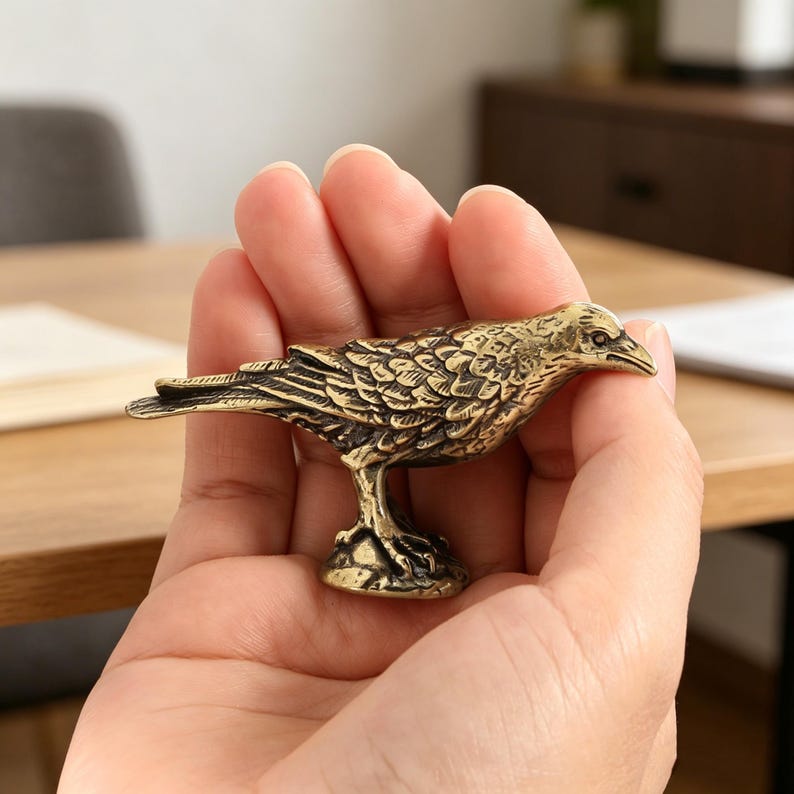 May include: A detailed brass raven figurine. The bird is perched on a small base, with intricate feather detailing. The raven is held in a person's hand, showcasing its size and craftsmanship. The figurine has a vintage, antique appearance.