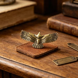 Peut inclure: Une figurine d'aigle en laiton doré avec des ailes déployées, montée sur une petite base carrée en cuir marron. L'aigle est de couleur dorée et la base en cuir est marron chaud. Le fond comprend des livres anciens et une surface en bois.