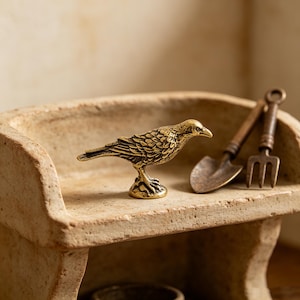 May include: A detailed brass bird figurine with textured feathers stands on a small pedestal. Beside it are miniature gardening tools: a shovel and a fork. The items rest on a weathered, light-brown, ceramic surface.