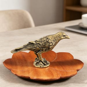 May include: A detailed brass bird figurine perched on a wooden, leaf-shaped tray. The bird has textured feathers and a sharp beak. The tray is a warm brown color, and the bird is a golden hue. The figurine is a decorative item.