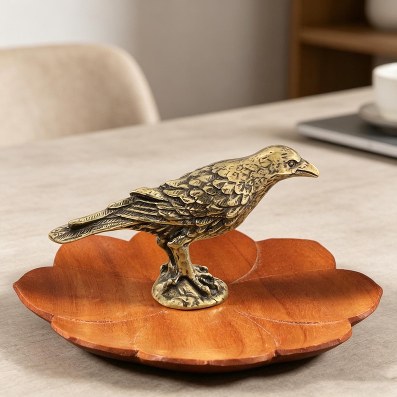 May include: A detailed brass bird figurine perched on a wooden, leaf-shaped tray. The bird has textured feathers and a sharp beak. The tray is a warm brown color, and the bird is a golden hue. The figurine is a decorative item.
