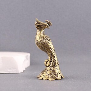 Puede incluir: Una peque&ntilde;a figura de lat&oacute;n de estilo antiguo que representa un f&eacute;nix de pie. El ave presenta detalles intrincados y texturizados de plumas en todo el cuerpo, una cabeza con cresta y una cola en forma de abanico que sirve de base.
