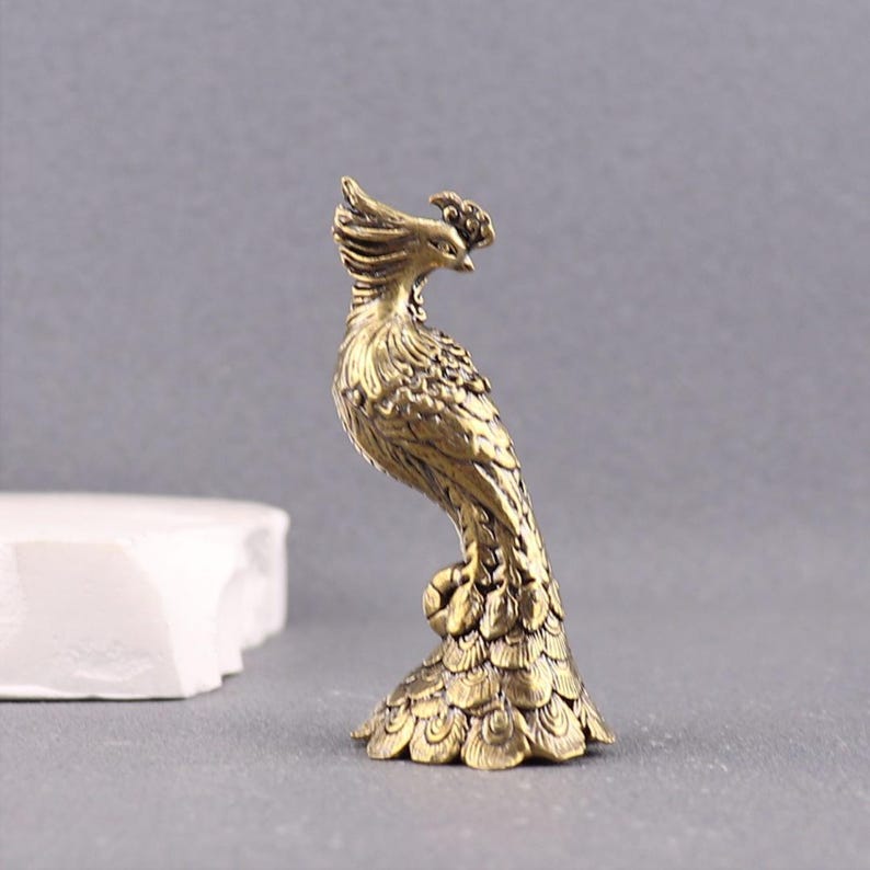 Puede incluir: Una peque&ntilde;a figura de lat&oacute;n de estilo antiguo que representa un f&eacute;nix de pie. El ave presenta detalles intrincados y texturizados de plumas en todo el cuerpo, una cabeza con cresta y una cola en forma de abanico que sirve de base.