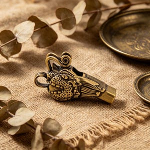 Könnte beinhalten: Eine detaillierte Messingpfeife mit einem verzierten Design mit einem stilisierten Gesicht und Schnörkeln. Die Pfeife hat eine warme Goldfarbe und liegt auf einer strukturierten, beigefarbenen Oberfläche. Zwei kleine Messingplatten befinden sich im Hintergrund.