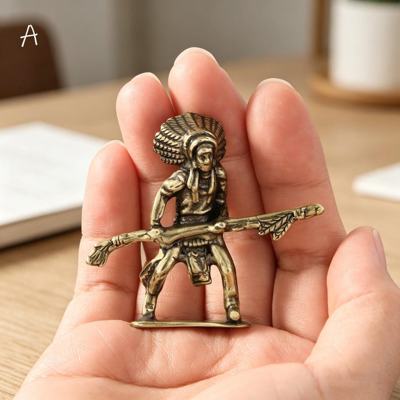 May include: A small, detailed brass figurine of a Native American figure holding a staff. The figure is wearing a headdress and is standing on a small base. The figurine is held in a person's hand.