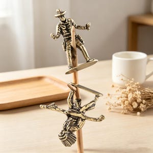 May include: Two bronze-colored cowboy figurines balancing on a wooden dowel. The top figurine is upright, wearing a hat and holding a gun. The bottom figurine is upside down, arms outstretched. The figurines are on a light-colored wooden surface.