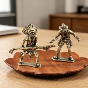 May include: Two brass figurines of a Native American and a cowboy stand on a wooden leaf-shaped tray. The Native American figure holds a staff, while the cowboy has a gun holster. The figurines are detailed and have a vintage aesthetic.