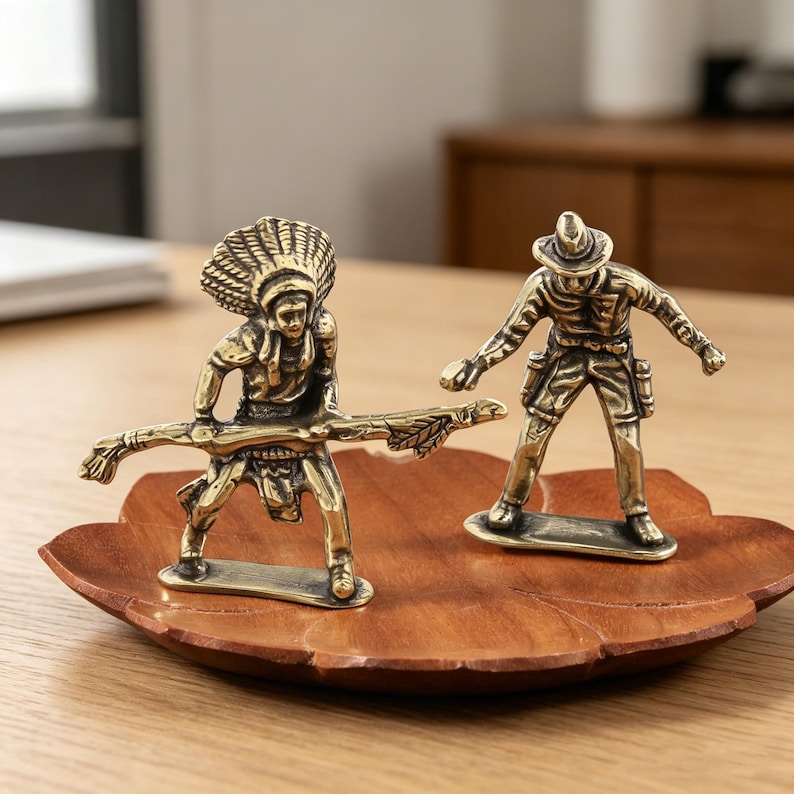 May include: Two brass figurines of a Native American and a cowboy stand on a wooden leaf-shaped tray. The Native American figure holds a staff, while the cowboy has a gun holster. The figurines are detailed and have a vintage aesthetic.