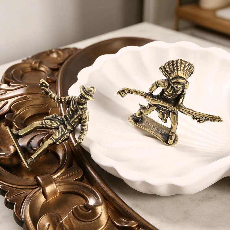 May include: Two bronze-colored figurines of people skateboarding. One is a cowboy, the other is an Indigenous person wearing a headdress. They are on a white seashell-shaped dish and a decorative brown tray.