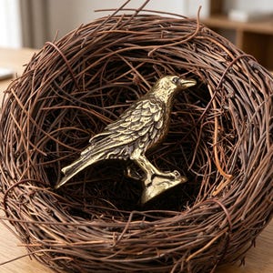 May include: A detailed brass raven figurine perched in a woven brown twig nest. The raven is a golden color with intricate feather detailing. The nest is round and made of intertwined twigs, creating a natural, rustic aesthetic.