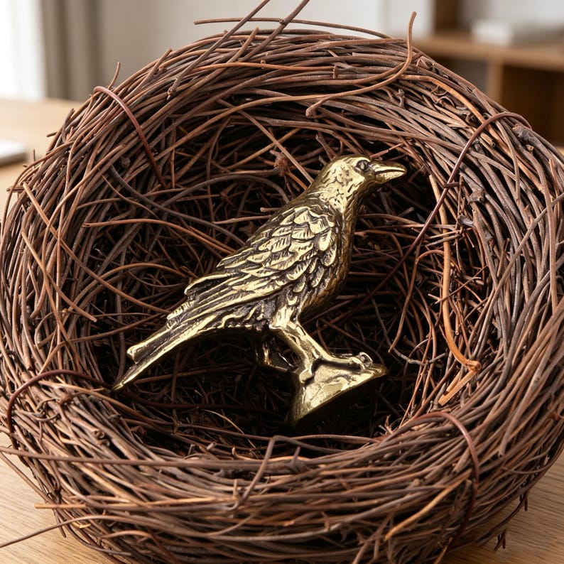 May include: A detailed brass raven figurine perched in a woven brown twig nest. The raven is a golden color with intricate feather detailing. The nest is round and made of intertwined twigs, creating a natural, rustic aesthetic.
