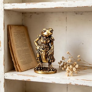 May include: A gold-toned metallic figurine of an anthropomorphic frog standing upright. The frog is depicted wearing ornate, detailed armor with intricate patterns on the shoulders and arms, and a draped cloth around its waist, set on a circular base.