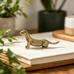 Peut inclure: Petite figurine de furet en laiton. Le furet est représenté debout, la tête légèrement tournée. La figurine présente des détails texturés et est posée sur une surface blanche. Des plantes vertes en arrière-plan.