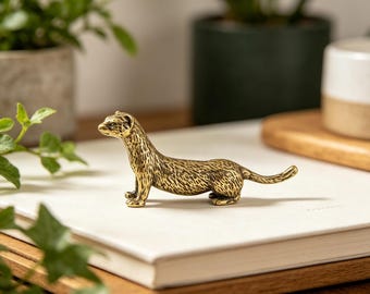 Puede incluir: Pequeña figura de hurón de latón detallada. El hurón está en posición de pie con la cabeza ligeramente girada. La figura tiene pelaje texturizado y está colocada sobre una superficie blanca. Hay vegetación verde en el fondo.