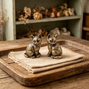 May include: Two bronze cat figurines sit on a burlap cloth inside a wooden tray. The cats are detailed with etched fur and alert expressions. The background includes a rustic shelf with dried flowers, creating a vintage aesthetic.