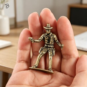 May include: A small, bronze-colored cowboy figurine. The figure is holding two guns and wearing a hat, jacket, and pants. The figurine is standing on a small base. The letter "B" is in the top left corner.