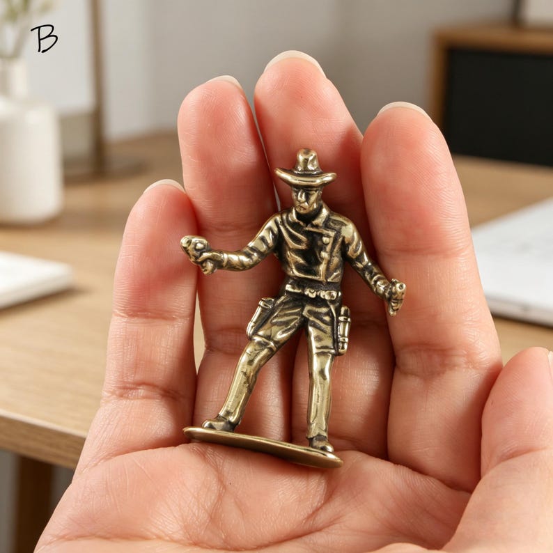 May include: A small, bronze-colored cowboy figurine. The figure is holding two guns and wearing a hat, jacket, and pants. The figurine is standing on a small base. The letter "B" is in the top left corner.