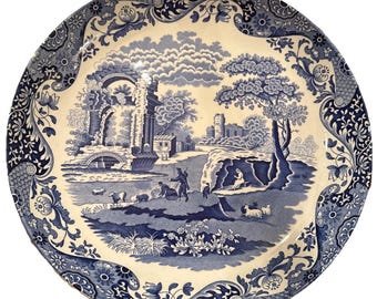 May include: A round, blue and white ceramic plate with a pastoral scene. The plate features a detailed landscape with figures, trees, and buildings. The rim has a decorative floral and scroll pattern. The plate is a classic piece of tableware.