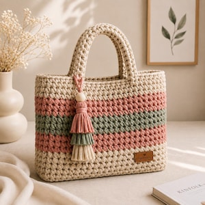 Könnte beinhalten: Eine rechteckige Häkeltasche mit horizontalen Streifen in Creme, Altrosa und Salbeigrün. Die Tasche hat zwei stabile Griffe und eine dekorative, dreistufige Quaste in passenden Farben an einer Seite. Ein kleines braunes Etikett trägt die Aufschrift Hand Made.