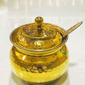 Op de afbeelding: Een goudkleurige metalen pot met een gehamerde textuur, voorzien van een bijpassend deksel met een centrale knop en een klein lepeltje dat erin rust. De houder heeft een afgerond lichaam en een gladde, reflecterende afwerking die het licht opvangt, waardoor heldere accenten op het metalen oppervlak ontstaan.