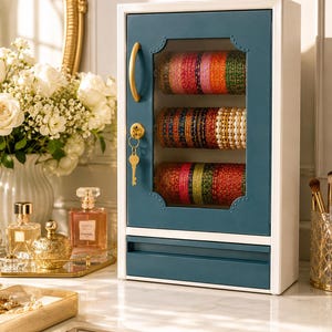 Op de afbeelding: Een rechthoekige sieradenvitrine voor aan de muur met een wit frame en een blauwgroene deur met een glazen venster. Binnenin houden drie horizontale stangen rijen kleurrijke armbanden vast. De deur heeft een gouden handgreep en slot.