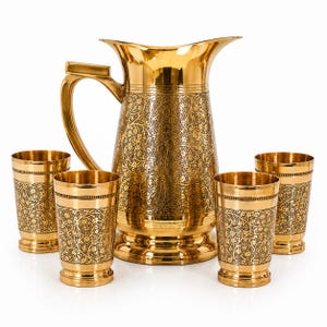 Op de afbeelding: Een goudkleurige metalen kan met een gebogen handvat en een bijpassende set van vier cilindrische bekers. Elk item is voorzien van een gedetailleerd, donker, geëtst bloemmotief over het oppervlak, met gladde, gepolijste gouden randen en voeten.