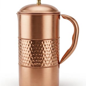 May include: A cylindrical copper pitcher with a brushed finish, featuring a central band of hammered texture bordered by beaded detailing. The pitcher includes a matching lid with a small brass-colored knob and a curved handle on the side.