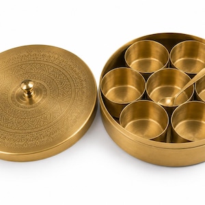 Op de afbeelding: Een ronde kruidendoos van messing met een bijpassend deksel voorzien van een ingewikkeld gegraveerd patroon en een centrale knop. De houder bevat zeven kleine, uitneembare messing bakjes in een cirkel, met een klein messing lepeltje erin.
