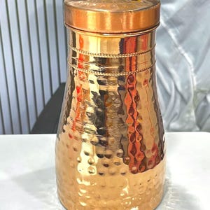 Könnte beinhalten: Eine zylindrische Wasserflasche aus Kupfer mit gehämmertem Textur-Finish. Die Flasche verfügt über einen glatten, flachen Deckel und ein dekoratives horizontales Band mit Perlenmuster nahe der Oberseite. Die Oberfläche ist poliert und reflektierend.