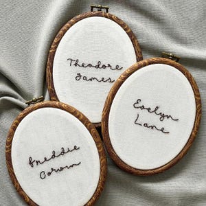 May include: Three oval embroidery hoops with faux wood grain frames containing white fabric. Each hoop features black hand-stitched lettering. The names embroidered are Theodore James, Evelyn Lane, and Freddie Corwin in a cursive script style.