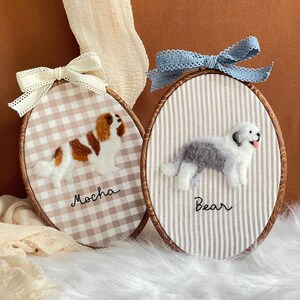 May include: Two oval embroidery hoops featuring needle-felted dogs on fabric. The left hoop has a brown and white dog with the name Mocha. The right hoop has a grey and white dog with the name Bear. Both hoops are topped with decorative bows.