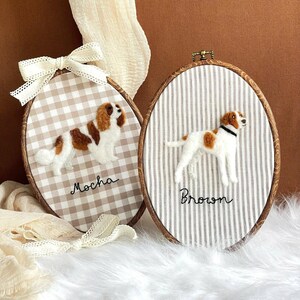 May include: Two oval embroidery hoops featuring needle-felted dogs. The left hoop has a beige gingham fabric with a brown and white dog labelled "Mocha" and a cream ribbon. The right hoop has a striped fabric with a white and brown dog labelled "Brown".