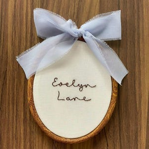 May include: An oval embroidery hoop with a wood grain finish containing white fabric. The name Evelyn Lane is stitched in dark brown thread in the centre. A light blue ribbon with frayed edges is tied in a bow at the top of the hoop.