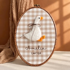 Può includere: Un telaio da ricamo ovale contenente un tessuto con motivo a quadretti beige e bianco. Un'oca in feltro bianco con becco e zampe arancioni è fissata sul tessuto, decorata con un sottile nastro marrone. Il nome Amelia Carter è ricamato sotto.