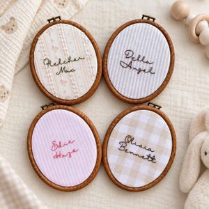 Op de afbeelding: Vier ovale houten borduurringen met stoffen inzetstukken met geborduurde namen. Links-boven bloemige strepen met 'Fletcher Mac', rechts-boven grijze strepen met 'Della Angel', links-onder roze strepen met 'Edie Haze' en rechts-onder beige ruitjespatroon met 'Olivia Bennett'.