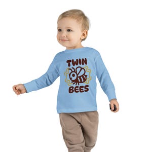 May include: A light blue long-sleeve shirt featuring a brown graphic of a bee with the text "TWIN BEES" in brown lettering. The design includes yellow zigzag lines surrounding the bee illustration, centred on the front of the shirt.