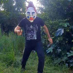 Peut inclure: Une personne portant un masque de clown avec un nez rouge, des yeux bleus et un chapeau marron. La personne porte un t-shirt noir avec un motif graphique et un pantalon noir, tenant une arme à feu dans une zone herbeuse.