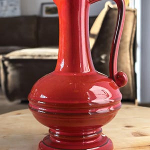 Könnte beinhalten: Glänzender roter Keramikkrug mit Henkel und bauchigem Boden. Der Krug hat einen Ausguss und enthält grüne Blätter. Er steht auf einem Holztisch. Ungefähr 25 cm hoch.