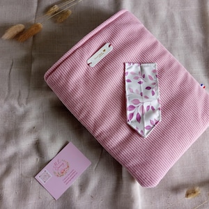 Peut inclure: Une housse d'ordinateur portable en velours côtelé rose avec une poche décorative en tissu blanc à motifs de feuilles violettes. Une petite étiquette est attachée à la housse. Une carte de visite avec un motif floral est également présente.