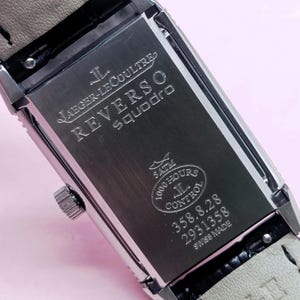 Pu&ograve; includere: Un orologio rettangolare in acciaio inossidabile con un cinturino in pelle grigio chiaro. Il quadrante dell'orologio presenta la scritta "JAEGER-LECOULTRE REVERSO Squadro" e altri dettagli. L'orologio &egrave; contrassegnato "SWISS MADE".