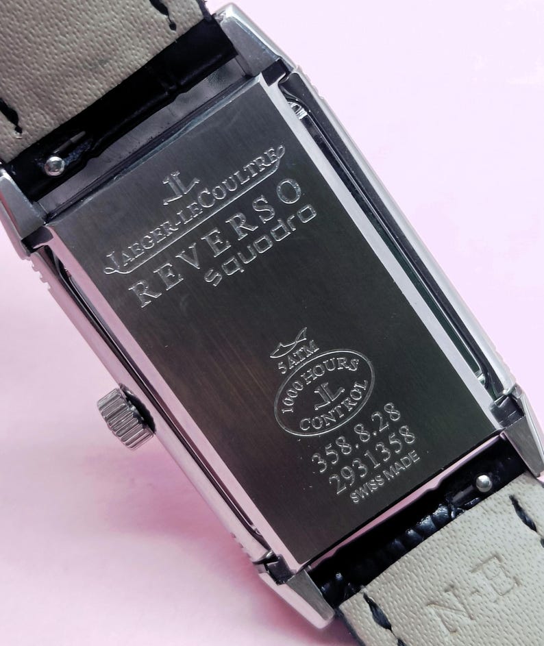 Pu&ograve; includere: Un orologio rettangolare in acciaio inossidabile con un cinturino in pelle grigio chiaro. Il quadrante dell'orologio presenta la scritta "JAEGER-LECOULTRE REVERSO Squadro" e altri dettagli. L'orologio &egrave; contrassegnato "SWISS MADE".
