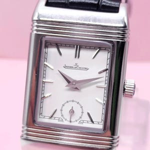 Pu&ograve; includere: Un orologio da polso rettangolare argentato con quadrante bianco, lancette nere e un piccolo quadrante dei secondi. L'orologio ha un cinturino in pelle nera e il marchio "Jaeger-LeCoultre" &egrave; visibile.