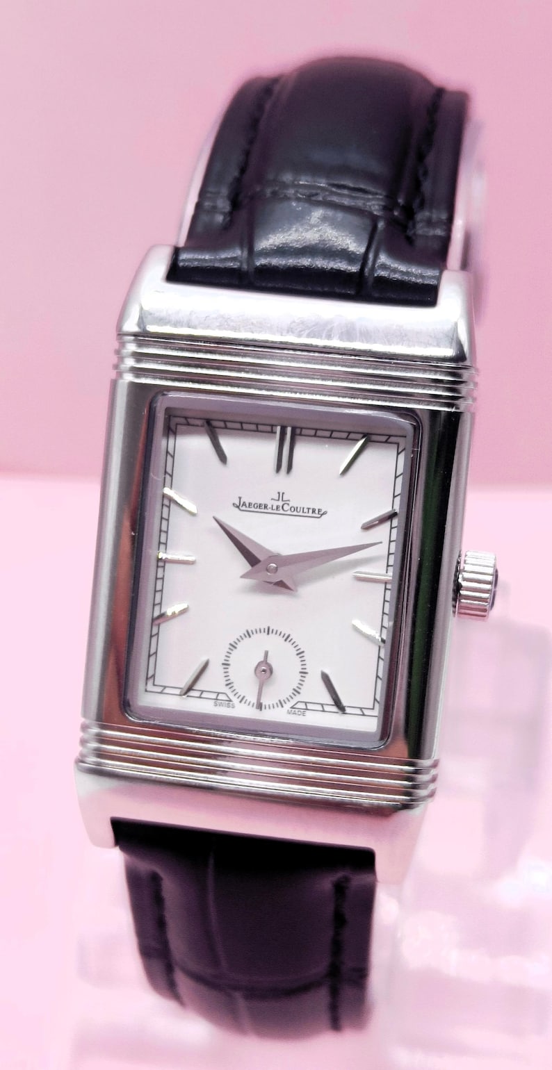 Pu&ograve; includere: Un orologio da polso rettangolare argentato con quadrante bianco, lancette nere e un piccolo quadrante dei secondi. L'orologio ha un cinturino in pelle nera e il marchio "Jaeger-LeCoultre" &egrave; visibile.