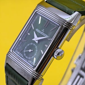May include: A rectangular silver-tone wristwatch with a dark green dial, silver hands, and silver hour markers. The watch features a small seconds sub-dial at the bottom, the Jaeger-LeCoultre logo, and a dark green leather strap with a textured finish.